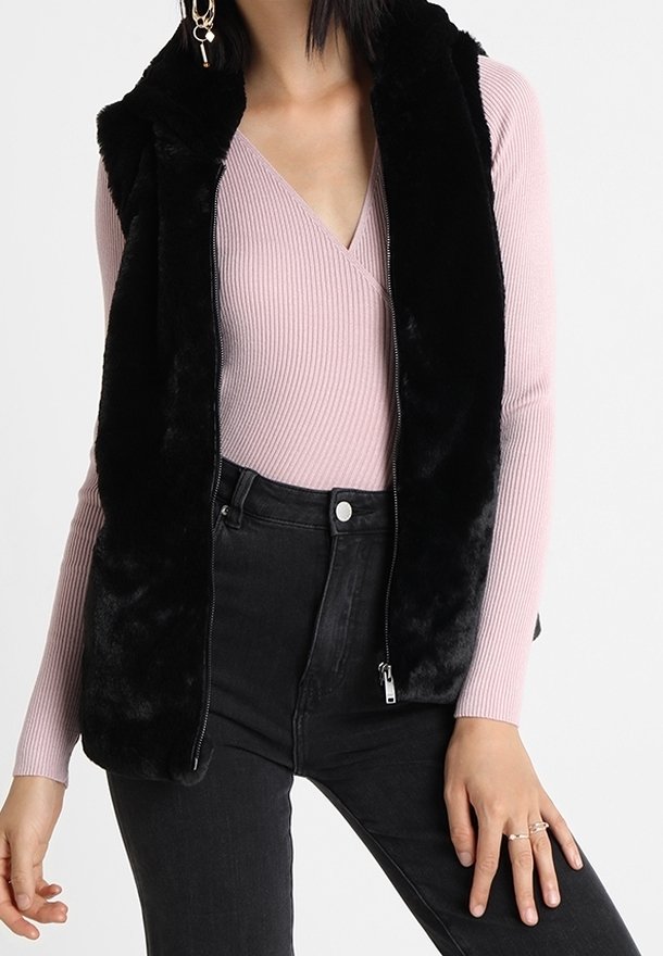 La femme porte un gilet sans manches en fausse fourrure noire sur un haut cache-cœur côtelé rose et un jean noir taille haute.
