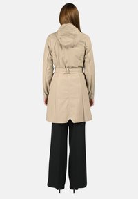 Μπεζ παλτό τρανς με κουκούλα, ζώνη στη μέση και ανοιχτή πλάτη. Φορεμένο πάνω από φαρδιά μαύρη παντελόνια, με λεία, ματ υφή.