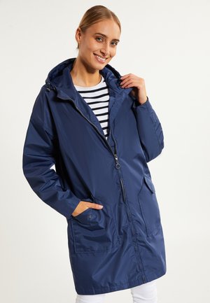 Lächelnde Frau trägt eine marineblaue Kapuzenregenjacke über einem schwarz-weiß gestreiften T-Shirt, mit einer Hand in der Tasche und der anderen, die den Kragen hält.