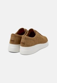 Brown suede low-top sneakers with white rubber outsole, featuring a smooth texture, stitched accents, and a circular logo on the heel.