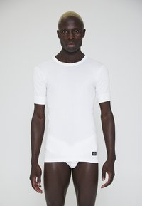 White short-sleeve fitted shirt, ribbed texture, round neckline, with a small black tag at the hem. Worn by a person.