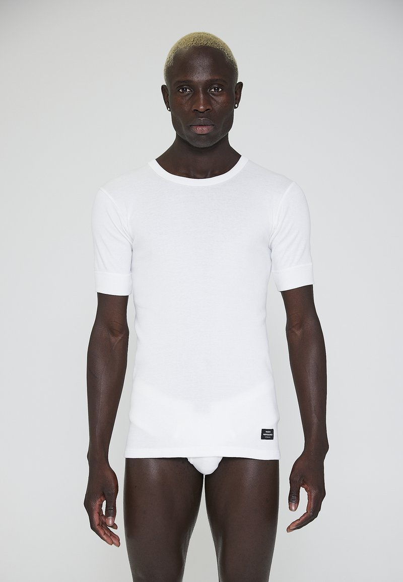 White short-sleeve fitted shirt, ribbed texture, round neckline, with a small black tag at the hem. Worn by a person.