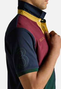 Krāsains polo krekls ar tumši zilu, bordo, dzeltenu un zaļu daļu; izteikta '3' izšūta uz piedurknes un zīmola nosaukums priekšā.