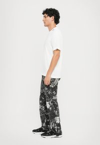 Camisa blanca de manga corta, pantalones negros con estampado de flores y calaveras, y zapatos deportivos negros, vistos de lado contra un fondo liso.