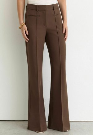 Vrouw draagt een hoge taillebruine wijde broek met frontplooien, gecombineerd met een wit topje en een gouden kettingarmband aan de linkerkant van de pols.