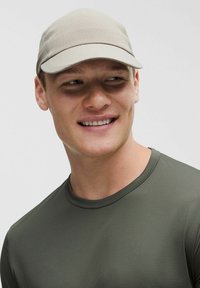 Casquette beige clair avec une texture côtelée, visière incurvée et détails de couture, portée avec une chemise ajustée verte olive.