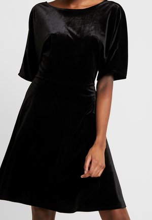 Czarna aksamitna sukienka do kolan z rękawami do łokci i okrągłym dekoltem, noszona przez osobę stojącą na jednolitym tle.