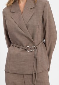 Blazer marron ajusté avec une finition texturée, doté d'une boucle en métal cintrant la taille. Col cranté et coupe décontractée.