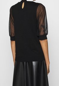 Svart stickad topp med genomskinliga puffärmar som har ett prickigt mönster, nyckelhål baktill och en avslappnad passform. Matchad med en svart läderskjorta.