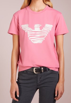 Osoba nosząca różowy T-shirt z białym stylizowanym logo orła, w połączeniu z ciemnymi jeansami i czarnym paskiem ze srebrną klamrą.