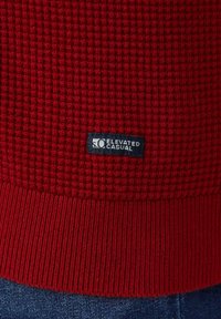 Roter, gestrickter Pullover mit strukturiertem Muster, gerippten Bündchen und einem marineblauen Label mit dem Schriftzug "Elevated Casual", das am Saum aufgenäht ist.