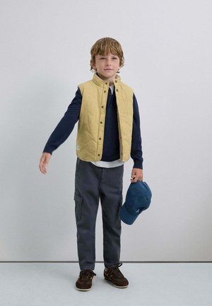 Chaleco acolchado amarillo sobre un suéter azul marino, pantalones cargo grises y zapatos marrones. Sosteniendo una gorra azul. Fondo simple, colores neutros.