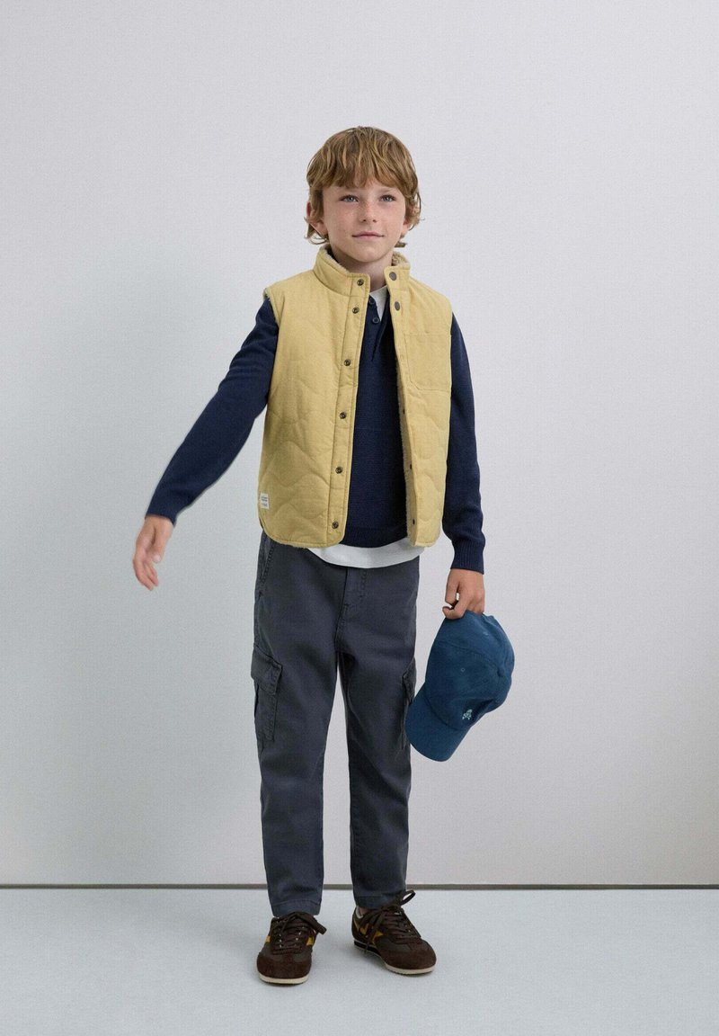 Gul quiltet vest over en marineblå sweater, grå cargo-bukser og brune sko. Holder en blå kasket. Simpel baggrund, neutrale farver.