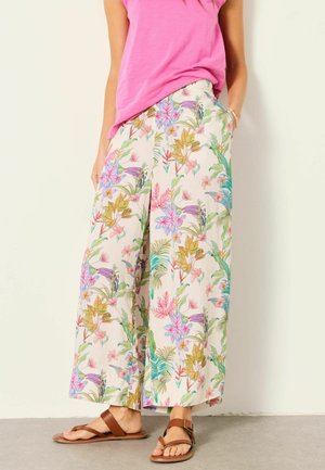 Femme portant un pantalon large crème avec imprimé floral coloré, un haut rose sans manches et des sandales marron, debout avec une main dans la poche.