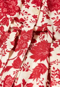 Tela con estampado floral rojo, con un lazo en la cintura y un botón blanco sobre un fondo crema, mostrando detalles de costura.