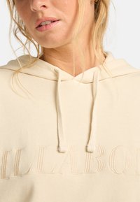 Beige hoodie met een getextureerd, geborduurd logo. Heeft een capuchon met trekkoord en zachte stof. Close-up van de halslijn en textuurdetails.