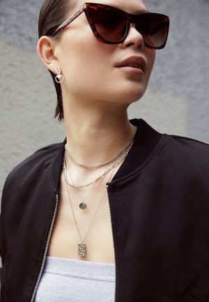 Óculos de sol em formato de cat-eye com padrão tartaruga, colares prateados em camadas e uma jaqueta bomber preta sobre uma blusa cinza claro. Close-up com detalhes texturizados.