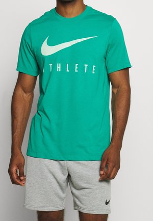 Mężczyzna w zielonej koszulce Nike z białym napisem "ATHLETE" oraz szarymi szortami Nike stoi na jednolitym tle.