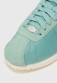 Aqua-blue sneaker featuring a mix of suede and nylon materials, textured laces, and a silver logo accent on the side. Rubber outsole.