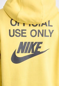 Gul huvtröja med dragsko i huvan. Har stor svart text med orden "ENDAST FÖR OFFICIELLT BRUK" och en framträdande svart Nike-logotyp.
