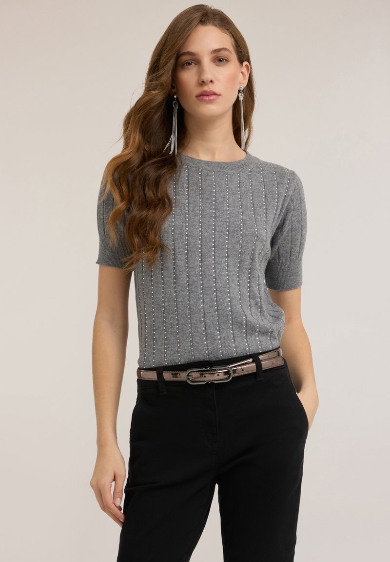 Suéter gris de manga corta tejido con canalé vertical y detalles de cuentas plateadas. Combinado con pantalones negros y un cinturón marrón brillante.