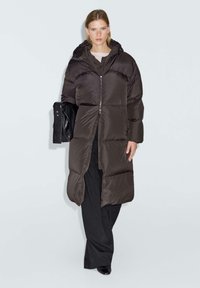 Donna che indossa un lungo cappotto imbottito marrone scuro con cappuccio, pantaloni neri a gamba larga, mentre tiene una borsa nera piegata su uno sfondo bianco puro.