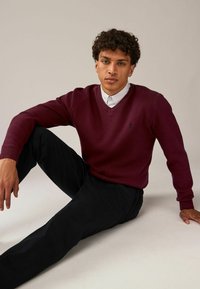 Pull en tricot bourgogne avec un col en V et des poignets côtelés, porté sur une chemise blanche à col, associé à un pantalon sombre.