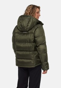 Vrouw met krullend haar in een olijfgroene gewatteerde jas met capuchon, gezien van achteren op een effen witte achtergrond.
