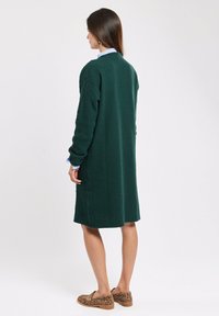 Long cardigan vert en tricot avec des motifs texturés, des fentes sur les côtés et un ourlet incurvé. Porté par-dessus une chemise à col bleu clair. Chaussures à imprimé léopard marron.