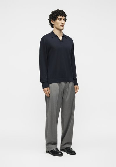 Νεαρός άνδρας όρθιος, φοράει μαύρο πουκάμισο με μακριά μανίκια και γιακά, γκρι παντελόνι με φαρδιά μπατζάκια και μαύρα επίσημα παπούτσια, με φόντο ένα απλό ανοιχτόχρωμο.