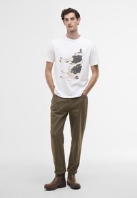 T-shirt branca com uma ilustração de uma vaca, combinada com calças bejes e botas de tornozelo castanhas. Design minimalista com um corte relaxado.