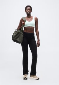 Αθλητική γυναίκα με μαύρο κολάν, ανοιχτοπράσινο αθλητικό σουτιέν και μπεζ αθλητικά παπούτσια, κρατώντας μια πράσινη τσάντα γυμναστηρίου στον ώμο της, σε λευκό φόντο.