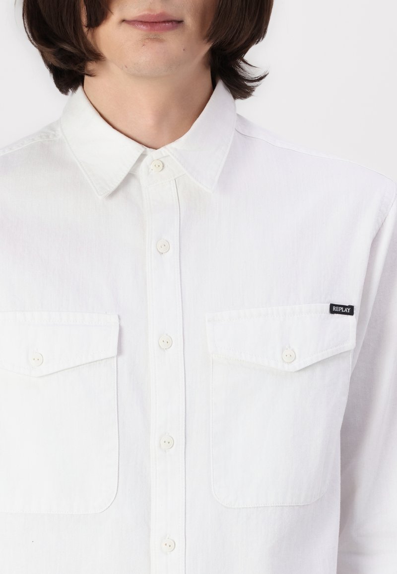 Man in een witte overhemd met knopen, voorzien van twee borstzakken, waarvan één met een klein zwart "REPLAY" label op de zakflap.