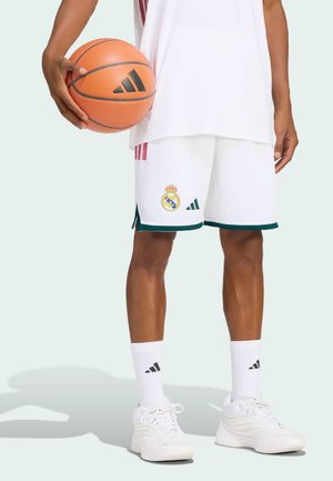 Person iført hvide Real Madrid basketballshorts, hvide sokker og sko, der holder en orange Adidas basketball mod en lys baggrund.