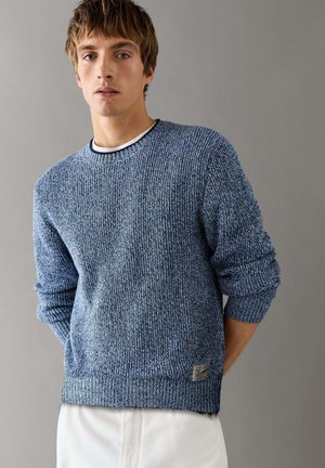 Jonge man met een blauwe gebreide trui met opgerolde mouwen en witte broek, staand tegen een effen grijze achtergrond.