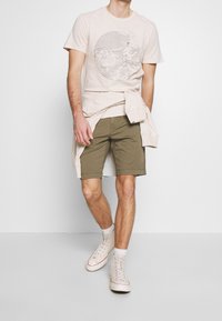 T-shirt beige con stampa di onde oceaniche, pantaloni corti verde oliva e scarpe da ginnastica bianche. Un maglione chiaro è legato in vita.