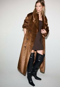 Cappotto in pelliccia sintetica marrone con ampi revers, bottoni doppi, un vestito aderente marrone e stivali neri in pelle al ginocchio.