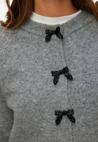 Szary sweter dziany z trzema ozdobnymi czarnymi kokardami na przodzie, z okrągłym dekoltem i fakturowaną tkaniną.