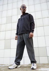 Felpa nera con cappuccio e tasca frontale, pantaloni grigi plissettati e sneakers bianche con dettagli neri. Il modello posa contro uno sfondo geometrico.
