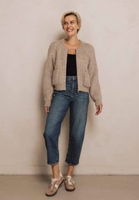 Hellbeige gestrickte Strickjacke mit Rippenstruktur, zwei Fronttaschen; kombiniert mit blauen, gekürzten Denim-Jeans und beigen Turnschuhen.