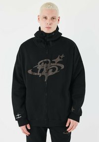 Sort zip-up sweatshirt med høj krave, stort logo i mørkebrun og subtile stjerneaccentuer. Stoffet ser blødt ud med en afslappet pasform.