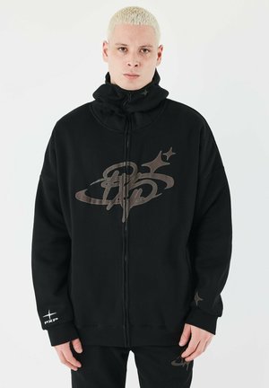 Sudadera negra con cremallera y alto cuello, gran logo en marrón oscuro y sutiles acentos de estrellas. El tejido parece suave y tiene un ajuste relajado.