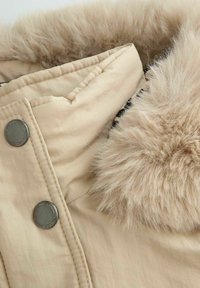 Beżowe okrycie wierzchnie z miękkim, sztucznym futerkiem na kołnierzu, z metalowymi guzikami i pikowanym wzorem, podkreślające ciepło i komfort.