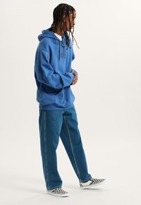 Sudadera azul con bolsillo frontal y logo, combinada con vaqueros de pierna ancha de color azul claro y zapatillas de lona de cuadros blancos y negros.