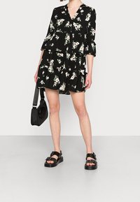 Robe noire à motif floral avec un design en étages, un décolleté en V et des manches à volants. Associée à des sandales noires et un petit sac noir.