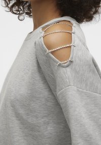 Felpa grigia chiara con design a spalla fredda, caratterizzata da dettagli in argento e texture a contrasto sulle maniche. Tessuto morbido, vestibilità rilassata.