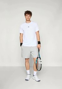 Fehér, rövid ujjú póló logóval, szürke rövidnadrág, fekete csuklópántok és fehér sportcipők. Teniszütőt tart.