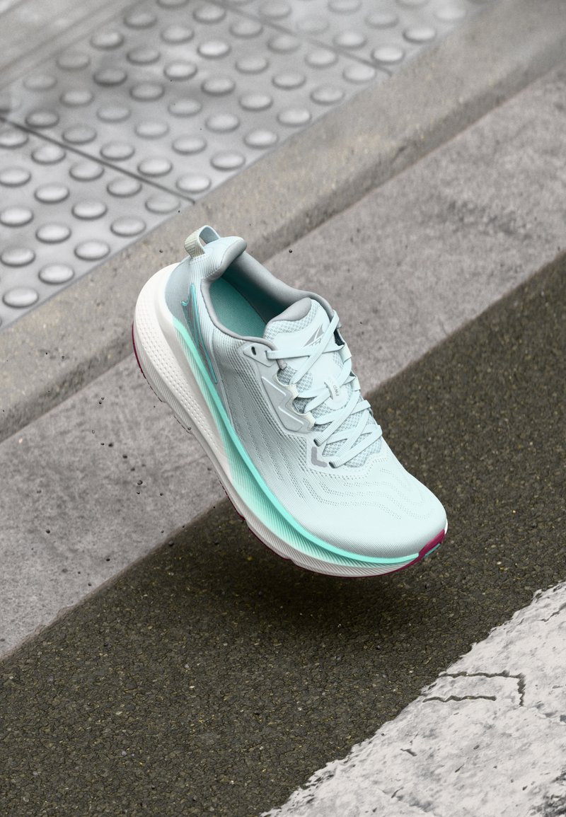 Light blue running shoe suspended above a street curb with tactile paving and asphalt road markings.
