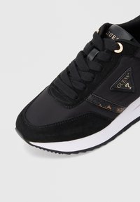 Czarny sneaker Guess z zamszowymi i skórzanymi detalami, czarnymi sznurowadłami, złotymi oczkami, naszywką z logo oraz białą gumową podeszwą.