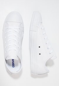 Paire de baskets blanches en cuir Converse basses, l'une vue de dessus montrant les lacets, l'autre de côté affichant la semelle et les trous de ventilation.